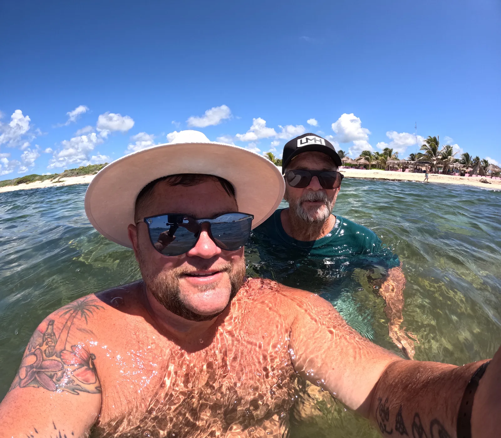 Chris and Randy, the son and dad who make Salt of the Page, taking a selfie waist-deep in clear turquoise water, with a white beach, palm trees and a blue sky behind them.